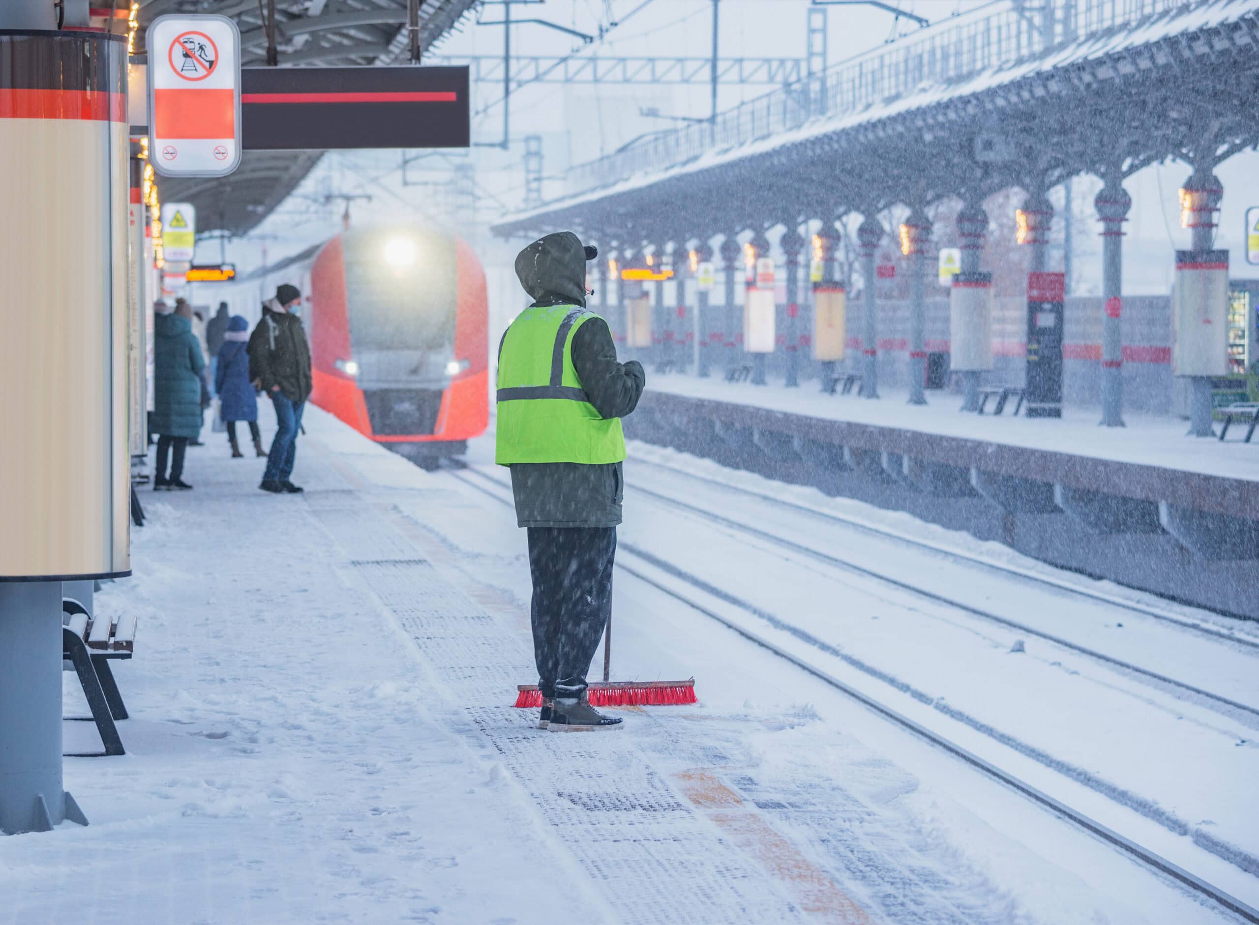 Ein Bahnsteig im Winter, bedeckt mit Schnee. Im Vordergrund steht eine Person mit einer neonfarbenen Warnweste und einem Besen, die den Schnee vom Bahnsteig fegt. Im Hintergrund nähert sich ein roter Zug dem Bahnhof, während andere Fahrgäste auf den Zug warten. Es schneit stark, und die Sicht ist durch den Schneefall eingeschränkt.